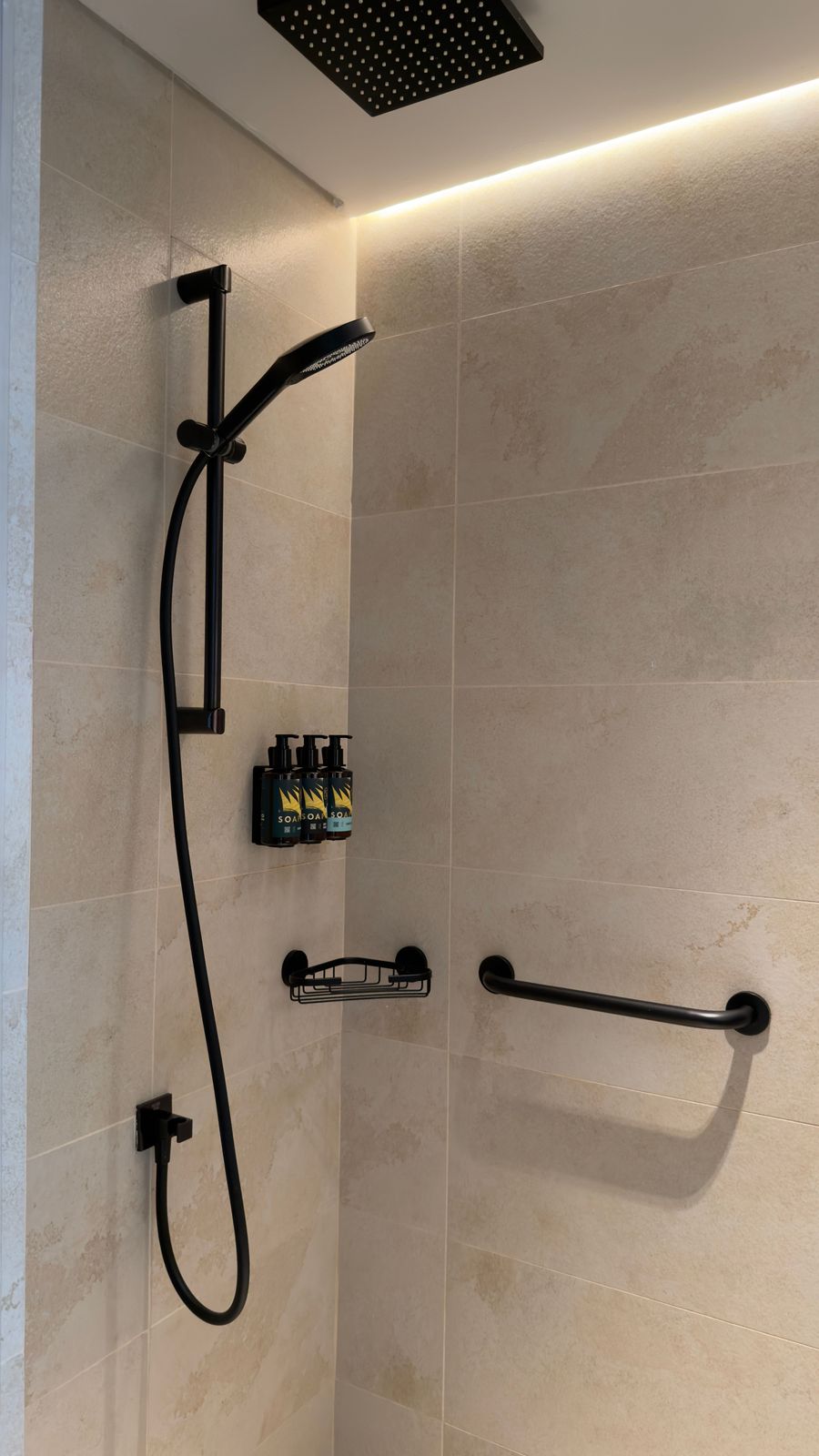 Matte black handheld shower on a slide rail in a warm-lit beige stone shower with toiletry holder and grab bar
