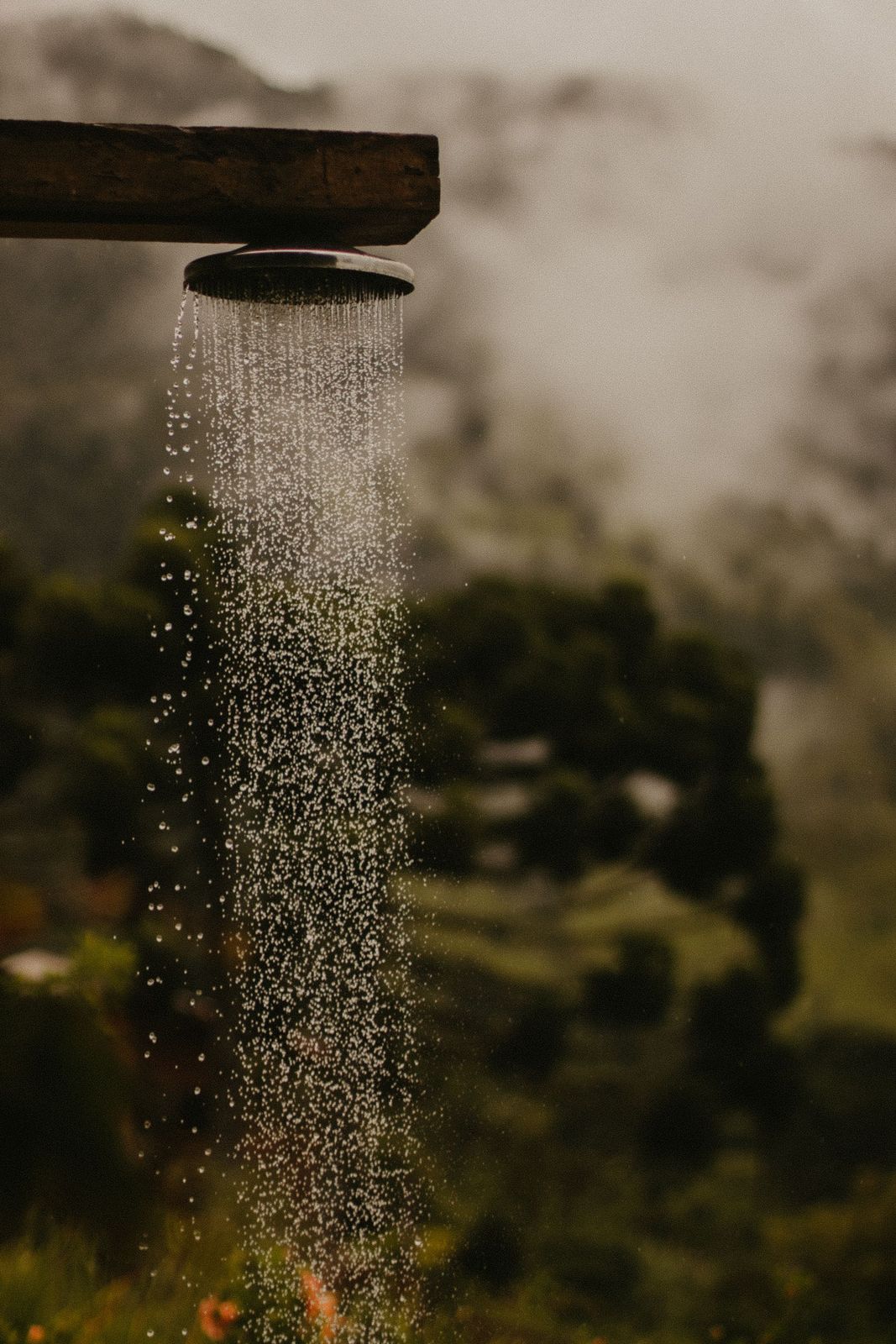 Round rain shower head mounted under a timber beam with water falling against misty green hills